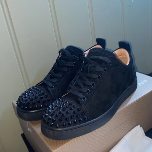 Svarta sneakers med nitar från Louboutin - Svarta sneakers i mocka från Louboutin med coola svarta nitar på tån. Skorna har rund tå, snörning och en platt sula. Insidan är ljusbeige och de har en lyxig vibe som sticker ut. Perfekta för dig som vill ha något extra till din outfit.