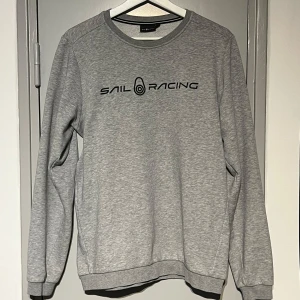 Grå sweatshirt från Sail Racing - Snygg grå sweatshirt från Sail Racing med svart logga tryckt över bröstet. Tröjan har rund halsringning, långa ärmar och ribbade muddar. Den är i storlek 176 och passar normalt till den. Perfekt för en chill och sportig look. Materialet är mjukt och skönt, troligtvis bomullsmix.