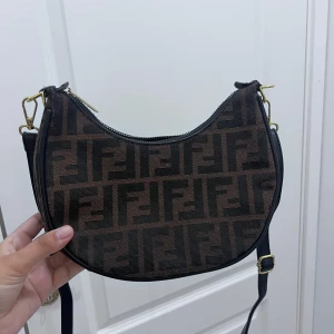 Fendi brun axelväska med logomönster - Säljer en brun axelväska från Fendi med klassiskt FF-logomönster i svart. Väskan har halvmåneform, justerbar axelrem och dragkedja upptill. På sidan sitter stora guldfärgade FENDI-bokstäver. Perfekt för dig som vill sticka ut med en ikonisk designeraccessoar.