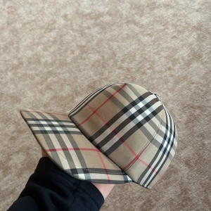Burberry rutig keps i beige och svart - Snygg keps från Burberry med klassiskt rutigt mönster i beige, svart, vitt och röda linjer. Justerbar snapback baktill och broderad Burberry London England-patch. Perfekt accessoar för att lyfta din outfit med lyxig streetkänsla.