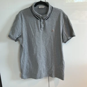 Grå Moncler pikétröja i bomull - Snygg grå pikétröja från Moncler med klassisk krage och svarta/vita ränder på krage och ärmslut. Broderad Moncler-logga på bröstet och knäppning framtill. Tillverkad i mjuk bomull, perfekt för en clean och stilren look. Änvänd fåtal gånger. Storlek L men passar ändå M, perfekt nu till sommaren. Köparen står för frakten.