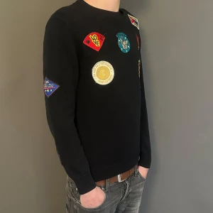 Svart Saint Laurent tröja med patches - Svart stickad tröja från Saint Laurent Paris med flera färgglada broderade patches på bröstet och ärmarna. Tröjan har rund halsringning och långa ärmar, perfekt för dig som vill sticka ut med en cool och unik look.