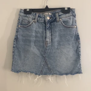 Ljusblå jeanskjol med fransig kant - Snygg ljusblå jeanskjol med rå, fransig nederkant och klassiska fickor fram och bak. Kjolen har bälteshällor, knapp och dragkedja framtill. Perfekt för dig som gillar en avslappnad och trendig stil. Står inte storlek men passar mig som har xxs-xs