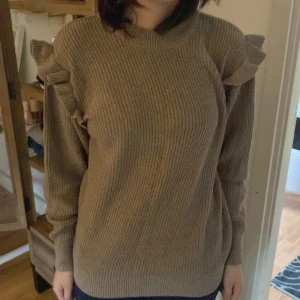 Beige stickad tröja med volangdetaljer - Säljer en beige stickad tröja med ribbad struktur och volangdetaljer på axlarna. Tröjan har rund halsringning och långa ärmar, perfekt för dig som gillar en söt och trendig look. Supermjuk och passar till många outfits!