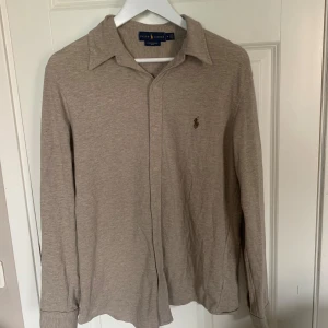 Beige skjorta från Ralph Lauren M - Stilren beige skjorta från Ralph Lauren i mjuk bomullskvalitet. Klassisk krage, långa ärmar och knäppning framtill. Diskret broderad logga på bröstet. Perfekt för dig som gillar en clean och avslappnad look.