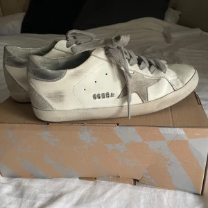 Golden Goose vita sneakers med silverdetaljer - Snygga vita sneakers från Golden Goose med grå snörning, silverfärgade detaljer på hälen och en ikonisk stjärna i mocka på sidan. 