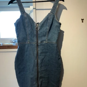 Jeansklänning med dragkedja från H&M - Snygg jeansklänning från H&M Divided i klassisk blå denim. Modellen är kort med breda axelband och markerad midja. Dragkedja framtill med stor ringdetalj ger en cool touch. Perfekt för dig som gillar en trendig och avslappnad stil.