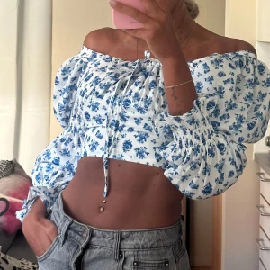 Blommig croppad blus med puffärm - Superfin croppad blus med blått blommönster på vit botten. Offshoulder-modell med puffiga långa ärmar och smock vid ärmslut. Söt knytning framtill och lätt volang vid axlarna. Perfekt för sommarens alla tillfällen!