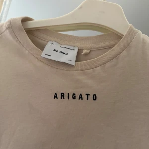 Beige t-shirt från Axel Arigato - Snygg beige t-shirt från Axel Arigato med svart broderad text på bröstet. Klassisk rund halsringning och avslappnad passform. 