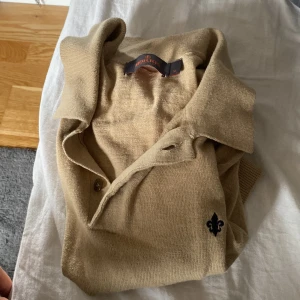 Beige pikétröja från Morris - Snygg beige pikétröja från Morris med klassisk krage och korta ärmar. Tröjan har knappar vid halsen och en broderad svart lilja på bröstet.       Knappt använd och inga defekter!     