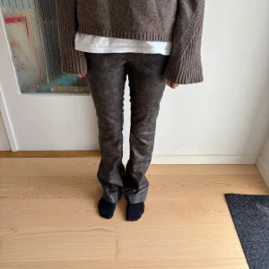 Bruna skinnbyxor från Monki - Snygga mörkbruna byxor i skinnimitation från Monki. Hög midja och raka ben med lätt bootcut. Byxorna har knapp och dragkedja framtill och är i ett mjukt syntetmaterial som ger en cool look.