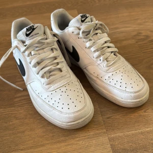 Nike Court Vision vita sneakers - Nike Court Vision sneakers i vitt skinn med svarta swoosh-loggor på sidorna. Klassisk låg modell med snörning och perforerade detaljer på tån för extra ventilation. Platt sula och bekväm mesh-fodring. Perfekta för en clean och sportig stil.