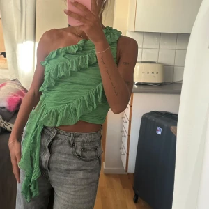 Grön oneshoulder volangtop - Säljer en grön, croppad topp med oneshoulder och volangdetaljer. Toppens tyg är lätt skrynklat och har en asymmetrisk form med volanger som går diagonalt över bröstet och ner längs sidan. 