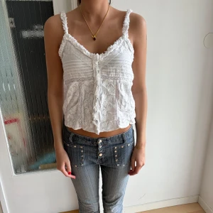 Vit spetsig linnetopp från Zara - Supersöt vit linnetopp från Zara med spetsdetaljer, volangkant och knappar framtill. Toppen har smala axelband och en lätt croppad passform. Perfekt för varma dagar när du vill ha en romantisk och chill vibe.