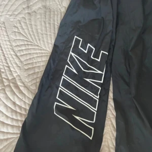 Byxor Nike - Svarta träningsbyxor från Nike med stort vitt logotryck längs ena benet. Byxorna har resår i midjan med snörning och är tillverkade i lätt syntetmaterial med meshfoder för extra komfort. Perfekta för gymmet eller chill.