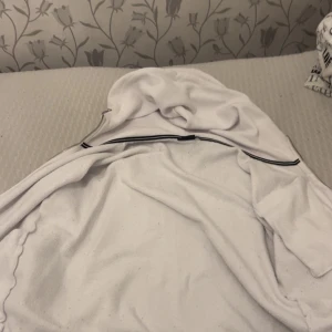 Vit hoodie från Sail Racing med dragkedja - Vit hoodie från Sail Racing med hel dragkedja och huva. Tröjan har en bröstficka med dragkedja och Sail Racing-logga på bröstet. Tillverkad i bomull och polyester, perfekt för chill dagar eller när du vill ha en clean look.