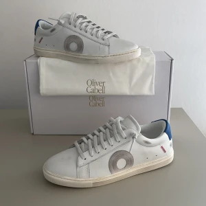 Oliver Cabell Low 1 COWBOY  - Snygga vita sneakers från Oliver Cabell med blå hälkappa och coola grå och röda detaljer på sidorna. Skorna har snörning, rund tå och är gjorda i skinn. Perfekta för dig som gillar stilrena men unika sneakers med en twist. Oliver cabellsen är sprillans nya och inte använda alls pågrund av att skorna inte passade mig 