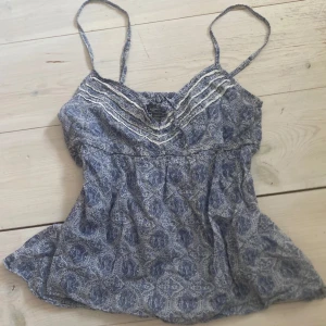 Blå mönstrad linnetopp från H&M - Säljer en blå och vit mönstrad linnetopp från H&M med tunna axelband och v-ringad detalj framtill. Toppen har ett bohemiskt paisleymönster och smockad rygg för extra komfort. Perfekt för varma dagar!