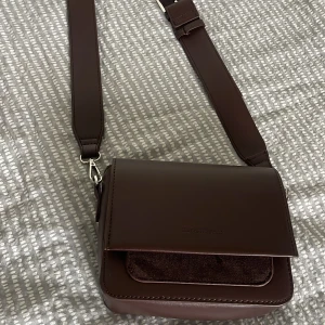 Brun axelväska från Carin Wester  - Snygg brun axelväska från Carin Wester i skinn med bred justerbar rem och magnetlås. Väskan har en ficka framtill i mocka och ett rymligt huvudfack med innerficka med dragkedja. Perfekt storlek för mobil, plånbok och småprylar.