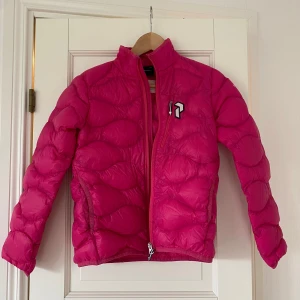 Rosa dunjacka Peak Performance 150 - Snygg rosa dunjacka från Peak Performance i storlek 150. Jackan har quiltad design, hög krage och dragkedja framtill. Perfekt för kyliga dagar och har en cool logga på bröstet. 