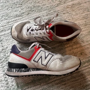 New Balance sneakers i vitt och rött -  New Balance sneakers med vit bas, grå mockadetaljer, röda och mörkblå inslag samt stora N-loggor på sidorna. 