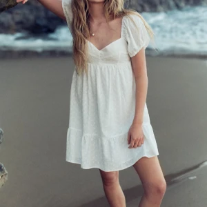 Vit kort sommarklänning med puffärm - Superfin vit kort klänning med puffärmar och v-ringning. Klänningen har broderade detaljer och volang nertill, vilket ger en romantisk vibe. Perfekt för varma dagar och har en luftig passform. Tillverkad i bomull för en skön känsla.