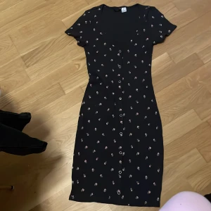 Svart blommig kortklänning H&M - Supersöt svart kortklänning från H&M med små rosa blommor, v-ringning och knappar framtill. Klänningen har korta ärmar och är gjord i ett mjukt, ribbat material som sitter snyggt på kroppen. Perfekt för dig som gillar en feminin och chill stil.