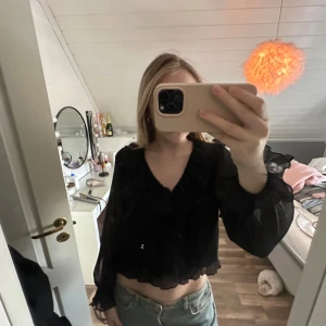 Svart volangblus med v-ringning - Säljer en svart blus med v-ringning och volangdetaljer. Blusen är croppad och har transparenta ärmar med lätt volym. Är i storlek m men sitter som en s. 