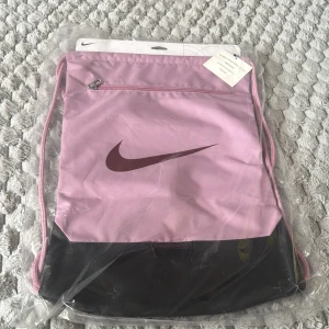 Nike rosa och svart gympåse - Säljer en snygg gympåse från Nike i ljusrosa och svart med mörklila swoosh-logga. Väskan har dragskostängning, en dragkedjeficka framtill och är tillverkad i slitstark polyester. Perfekt för träning eller att bära med sig grejer till skolan.