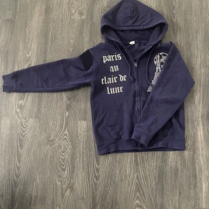 Mörkblå hoodie från bling&lucid - Mörkblå hoodie från bling&lucid med dragkedja och huva. Storlek s/smal. Framsidan har texten 'paris au clair de lune' och en glittrig Eiffeltorn-design i stendiamanter. Perfekt för dig som gillar Paris-vibes och snygga detaljer. Några pärlor har försvunnit men funkar ändå riktigt bra