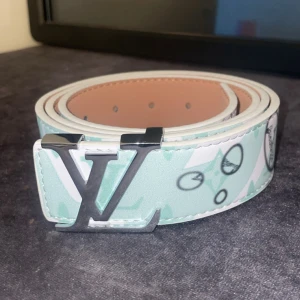 Louis Vuitton mintgrönt bälte med LV-spänne - Snyggt bälte från Louis Vuitton i mintgrönt och vitt med ikoniskt LV-mönster och silverfärgat LV-spänne. Bältet har en stilren design med grafiska detaljer och är gjort i skinn. Perfekt för att lyfta din outfit med en exklusiv touch.