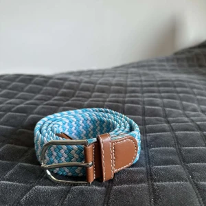 Blått och vitt flätat bälte med skinn - Snyggt flätat bälte i blått och vitt med detaljer i brunt skinn och silvrigt spänne. Bältet har en cool, randig look och passar perfekt till jeans eller shorts. Skinnet ger en schysst kontrast mot det färgglada bandet.