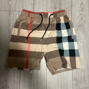 Burberry rutig badshorts med snörning - Snygga badshorts från Burberry med klassiskt rutigt mönster i beige, brunt, svart, vitt och blått samt röda detaljer. Shortsen har elastisk midja med svart snörning och sidofickor. Perfekta för sommaren och strandhäng.
