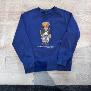 Blå sweatshirt Polo Bear Ralph Lauren - Blå sweatshirt från Polo Ralph Lauren med ikoniska Polo Bear-trycket på bröstet. Tröjan har rund hals, långa ärmar och ribbade muddar. Perfekt för dig som gillar klass, fashion och bekvämlighet.