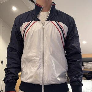 D&G Sport vindjacka i blått och vitt - Snygg vindjacka från D&G Sport i blått och vitt med röda och marinblå ränder på bröstet. Storlek 48 (s-m) passar bättre S. Nypris: 4500kr. 