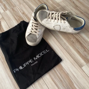 Philippe Model vita sneakers strl 40 - Snygga vita Philippe Model sneakers med grå mockadetaljer på tån och blå hälkappa. Klassisk låg modell med snörning och skinnmaterial. Sulan har coolt Philippe Model-tryck undertill. Perfekta för dig som gillar stilrena och trendiga sneakers.