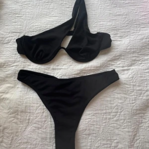 Svart bikini med asymmetrisk topp - Snygg svart bikini med ribbad struktur. Toppen har en unik asymmetrisk design med en axelrem och bygel för extra stöd. Underdelen är high-cut med rynkade detaljer bak för en trendig look. Perfekt för stranden eller poolen i sommar.