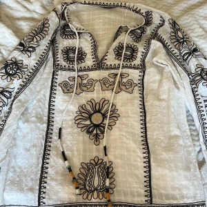  kort klänning med broderier från Zara🍓🫶🏼 - Säljer en superfin vit kort klänning från Zara med detaljerade svarta broderier. Den har en v-ringad design med snörning och pärlor i svart, vitt och orange. Perfekt inför sommar med sina långa ärmar och luftiga material. 🙌🏼🍓