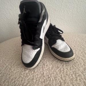 Nike Air Jordan 1 Low svart/vit 37,5 - Snygga Nike Air Jordan 1 Low sneakers i svart och vitt med grå detaljer. Klassisk siluett med svart snörning, perforerad tå och ikonisk swoosh på sidan. Skorna är i skinn och har en platt sula och rund tå. Perfekta för dig som gillar streetstyle. Lite nopprig på hälarna.