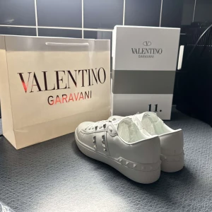 Valentino Rockstud vita sneakers - Snygga vita Valentino Rockstud sneakers i skinn med ikoniska silverfärgade nitar längs sidorna och på tungan. Klassisk rund tå, platt sula och vita snören. Perfekta för dig som vill sticka ut med en lyxig och edgy vibe.