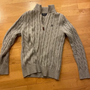 Grå kabelstickad tröja från Polo Ralph Lauren - Skitsnygg grå kabelstickad tröja från Polo Ralph Lauren med dragkedja vid halsen och broderad logga på bröstet. Riktigt stilren och passar till allt imellan vardagliga outfits till mer formella tillfellen. Kan shippa direkt. För mer frågor skicka ett meddelande!