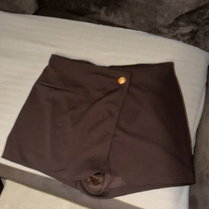 Brun omlottkjol från H&M - Mörkbrun kort shorts kjolfrån H&M med en snygg knappdetalj i midjan och en  ficka framtill. Kjolen har inbyggda shorts under för extra komfort och är tillverkad i ett mjukt, stretchigt material. Perfekt för dig som gillar stilrena och enkla plagg. Säljer då jag köpte fel storlek