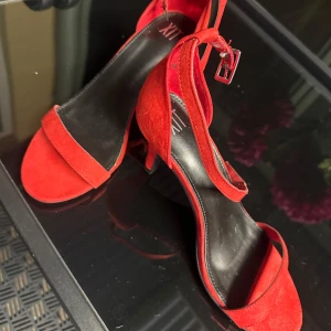 Röda sandaler med klack och ankelrem - Snygga röda sandaler från XIT med öppen tå, smal ankelrem och låg klack. Skorna har en stilren design i mocka och svart innersula, perfekta för att lyfta din outfit med en färgklick.