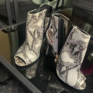 Ormskinnsmönstrade stövletter med klack - Snygga stövletter med ormskinnsmönster i svart, grått och beige. Skorna har öppen tå, hög klack och dragkedja på sidan. Perfekta för dig som vill sticka ut med en edgy och trendig look.