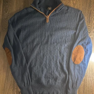 Blå kabelstickad tröja från Boggi - Mörkblå kabelstickad tröja från Boggi Milano i kashmir med half zip och snygga mockadetaljer vid dragkedjan och på armbågarna. Tröjan har lång ärm och ribbade muddar, perfekt för en stilren och chill look.