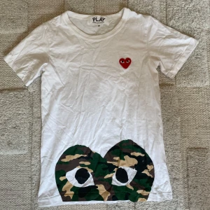 Vit Comme des Garçons Play t-shirt - Vit t-shirt från Comme des Garçons Play med klassiskt rött hjärta på bröstet och stor camo-ögon-grafik längst ner. Skön passform och tillverkad i mjuk bomull. Perfekt statement-plagg för dig som gillar streetstyle. Storleken är S men sitter lite mer som XS