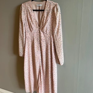 Ljusrosa blommig klänning från Adoore - Supersöt ljusrosa klänning från Adoore med små blommor, v-ringning och knappar hela vägen framtill. Långärmad modell med lätt markerad midja och flowig kjol. Perfekt för vår och sommar när du vill känna dig extra fin.