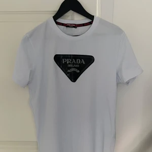 Ljusblå Prada t-shirt med logga - Snygg ljusblå t-shirt från Prada med klassisk svart logga framtill. Storlek M, liten i storleken så passar även S.                                                                   Helt ny!                                                        