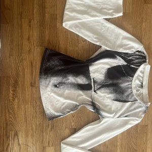 Vit långärmad topp med svart print - Säljer en vit långärmad topp med ett coolt svart grafiskt print framtill. Tröjan har rund halsringning och är figurnära i modellen. Materialet känns mjukt och stretchigt, perfekt för dig som gillar en stilren men edgy look.