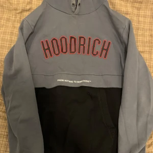 Hoodrich hoodie i grått och svart - Snygg hoodie från Hoodrich med grå överdel och svart nederdel. Stor röd logga framtill och texten 'From Nothing To Something' under. Klassisk huva och magficka, perfekt för en streetwear-look.